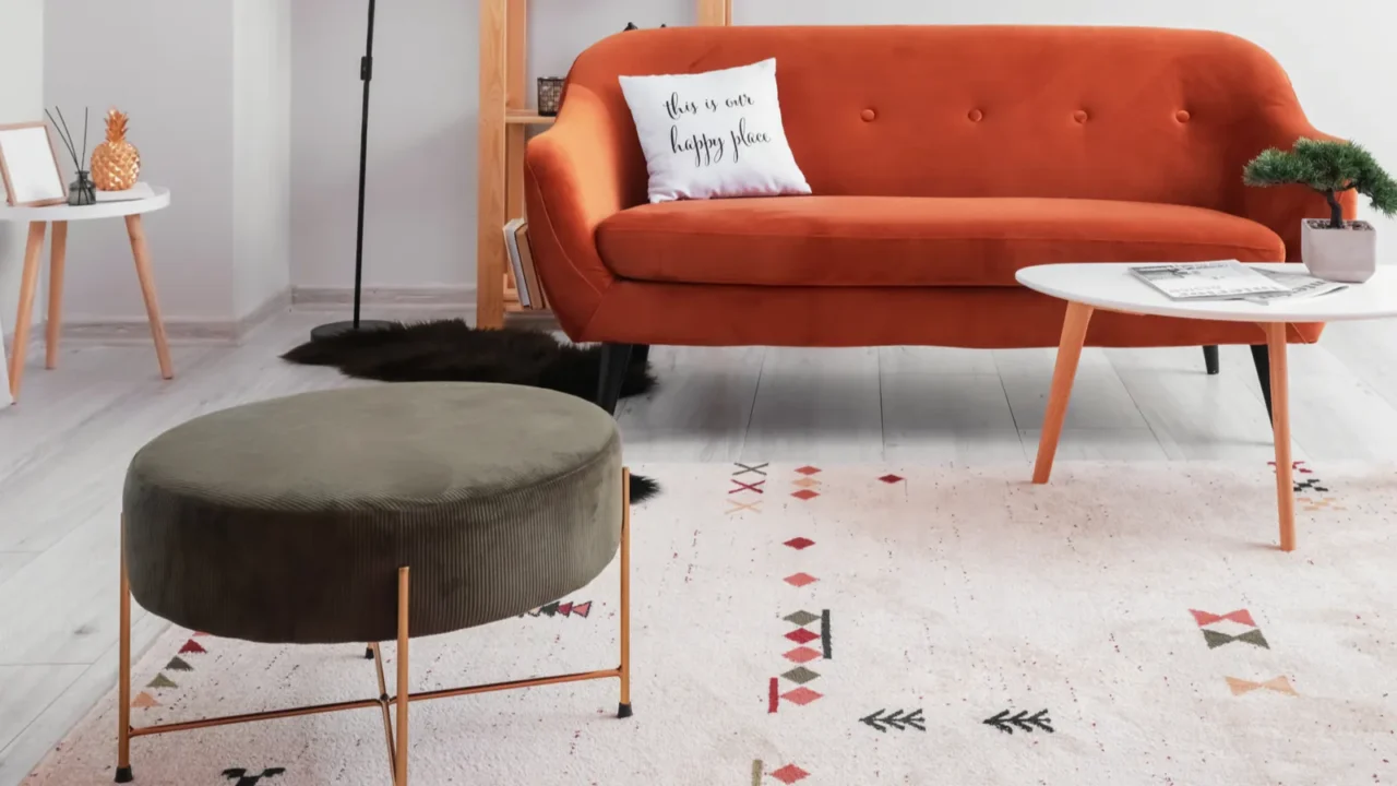 A living room interior with red sofa, coffee table with small potted bonsai, ottoman with gold frame, and a rug.