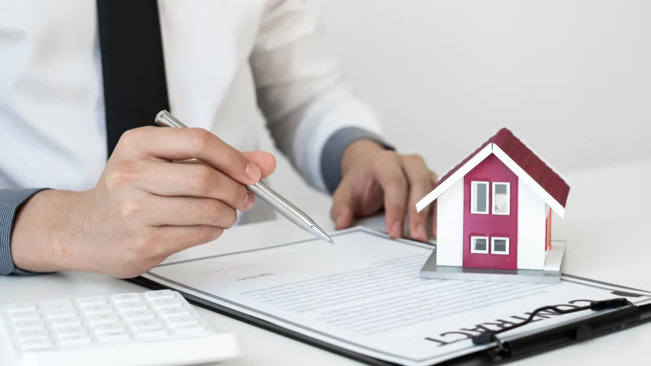 Person signing a real estate contract next to a small model house.