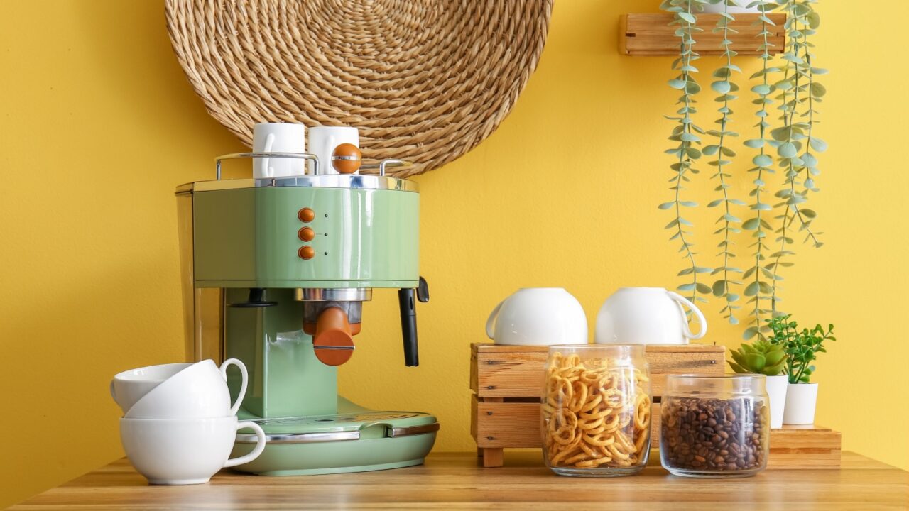 A coffee nook in a kitchen with green espresso machine with white cups stacked on top and jars of snacks beside it.