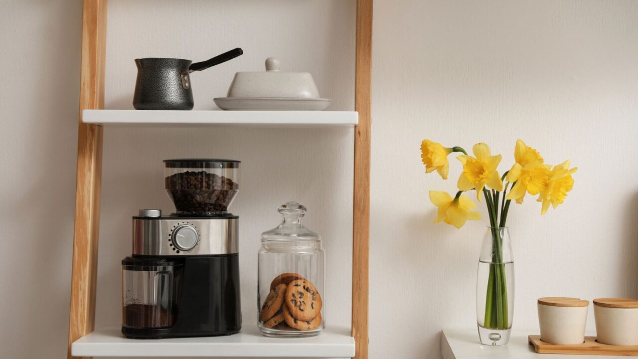 A shelf with a coffee grinder, kettle, cookies in a jar, and a vase of yellow flowers.