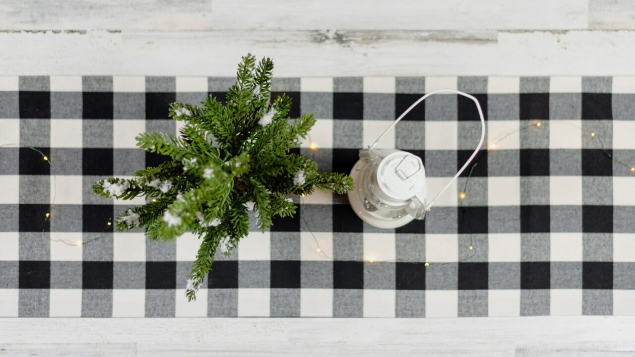 An overhead view of a small green plant beside a lantern on a black and white checkered tablecloth.