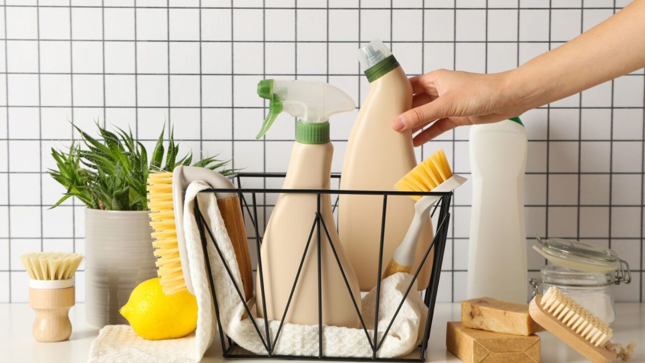 A variety of cleaning supplies organized in a basket on a tiled countertop with a plant and a lemon.