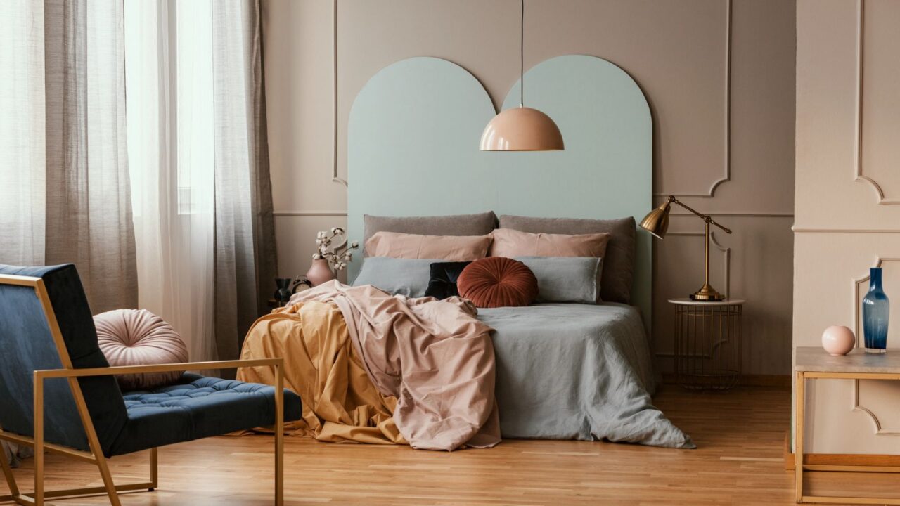 A stylish bedroom with a unique two-tone headboard, layered blankets, and a modern chair near large windows.