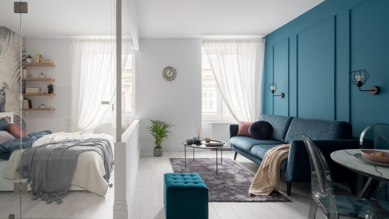 A small living space with a blue accent wall, a gray sofa, a round coffee table, and a cozy bed area separated by glass walls.