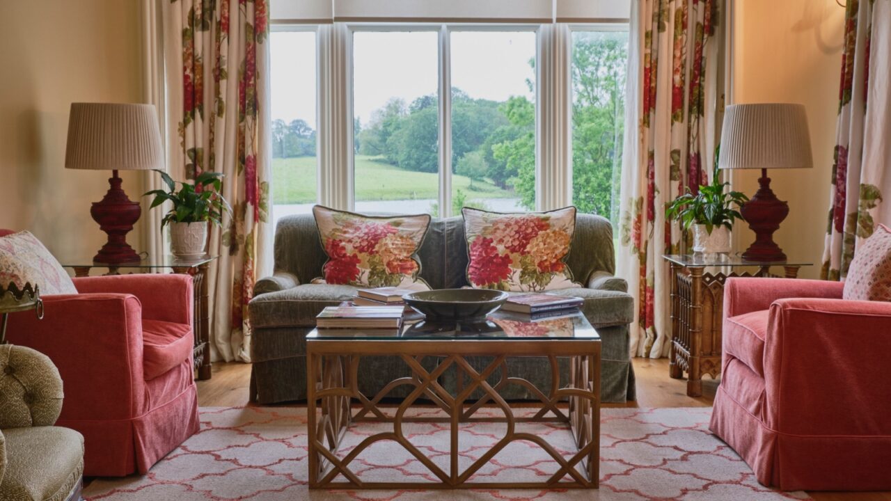 A living room interior with cozy sofas, throw pillows, coffee table, rug, and floral curtains on the windows.