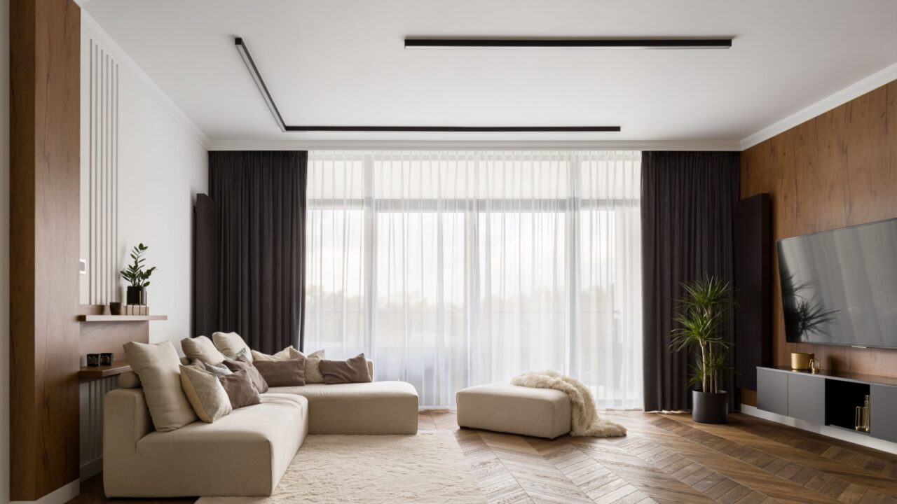 A modern living room with a beige sectional sofa, ottoman, large flat-screen TV, wooden accent wall, and floor-to-ceiling windows.