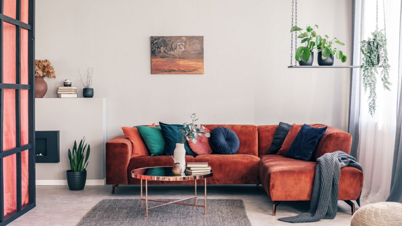 A cozy living room with a burnt orange sectional sofa, colorful cushions, a round coffee table, hanging plants, and a wall painting.