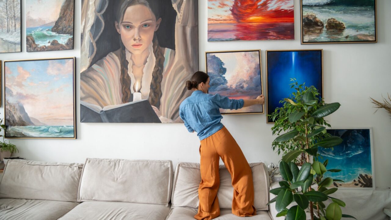 A woman adjusting artwork on a wall featuring several colorful paintings, situated in a modern, airy living room with a plush sofa and a potted plant nearby.