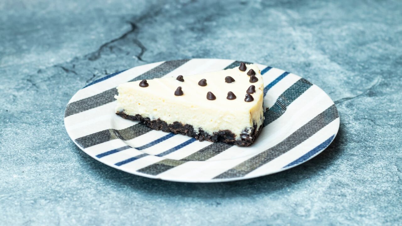 A slice of cheesecake with chocolate chips on a striped plate against a textured background.