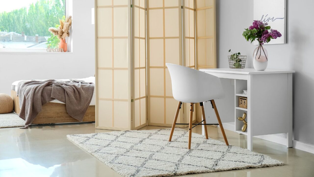 A bedroom interior with a bed, folding screen, a cozy rug, and a workspace with white table, chair, and a poster on the wall.