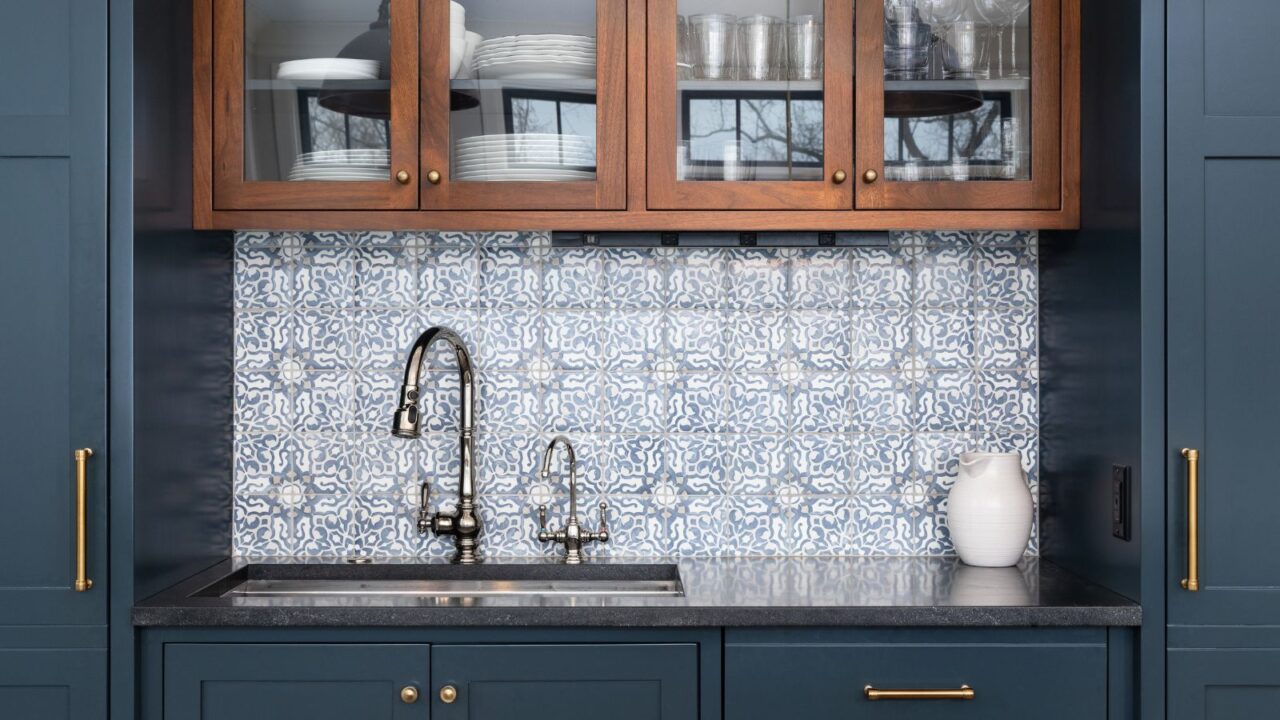 A kitchen interior with patterned tiles backsplash, chrome faucet, granite countertop, and blue and wood cabinetry.