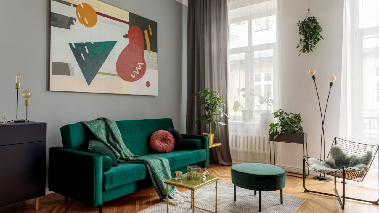 A living room interior with velvet sofa, coffee table, plants, rug, hanging planter, and abstract artwork on the wall.