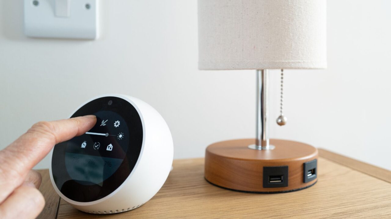 A person using a smart home hub device placed next to the lamp on a wooden table.