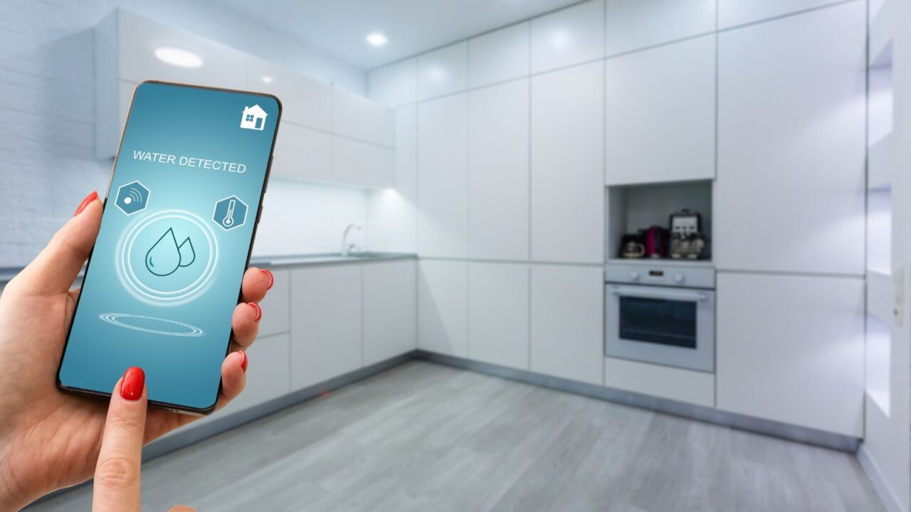 A woman holding a smartphone showing a water leak detector alert in a modern kitchen.