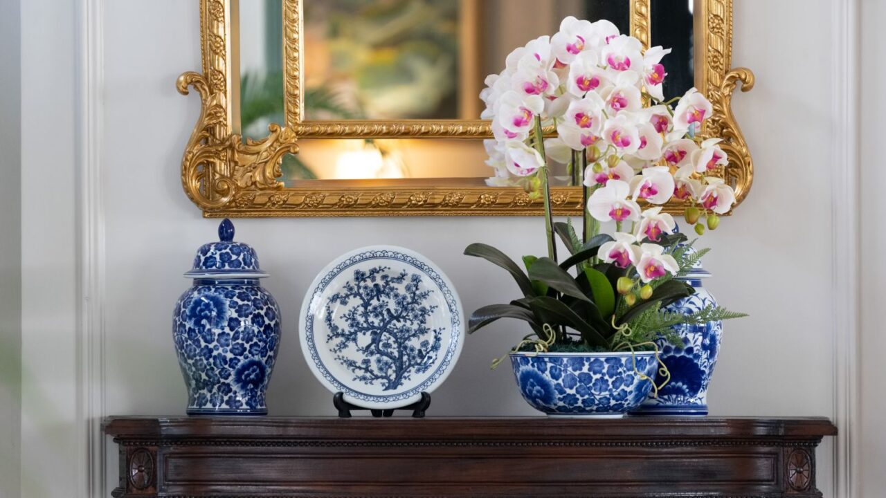 A blue paint porcelain collection with orchid vase on a dark wooden table with golden mirror on white wall.