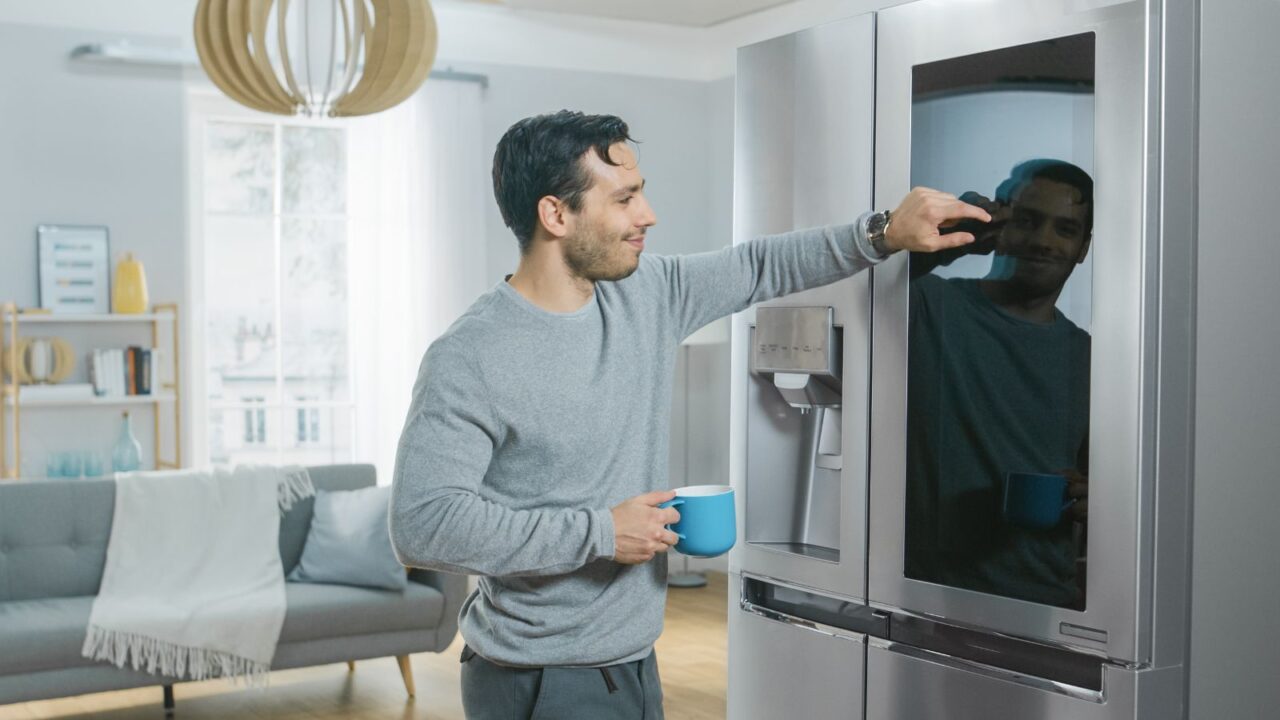 A man standing in front of a smart refrigerator while holding a blue mug.