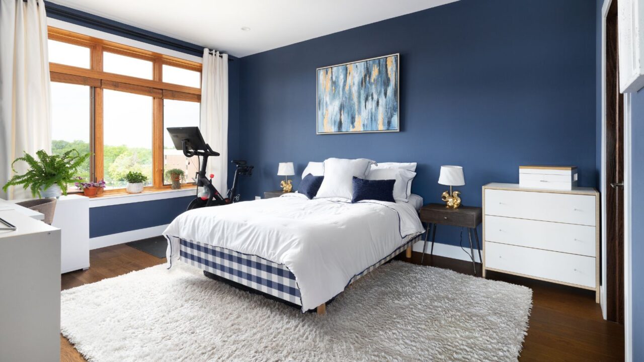 A stylish bedroom interior with a bed, plush rug, plants, curtains, nightstand, and framed artwork on the wall.