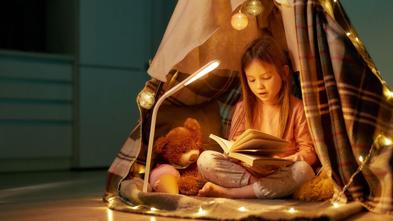 A girl reading a book in a cozy reading nook with a canopy, string lights, and stuff toys.