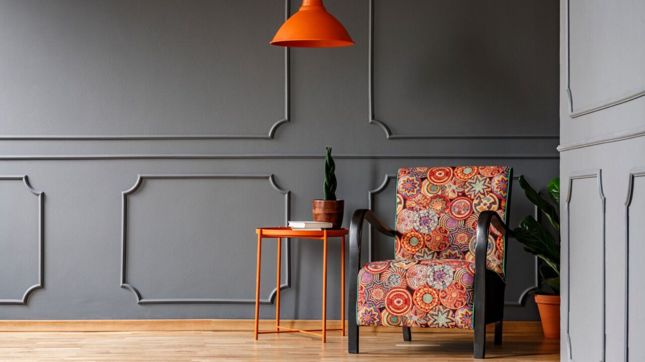 A living room interior with a boho style armchair, plant pot, and orange coffee table in front of a gray wall.