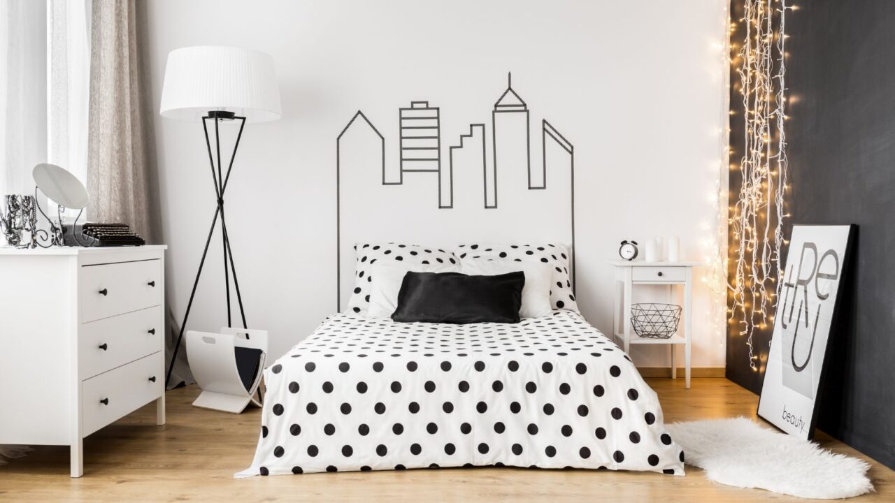 A bedroom interior with a cozy bed with black and white polka dots bedding, nightstand, poster, rug, and floor lamp.