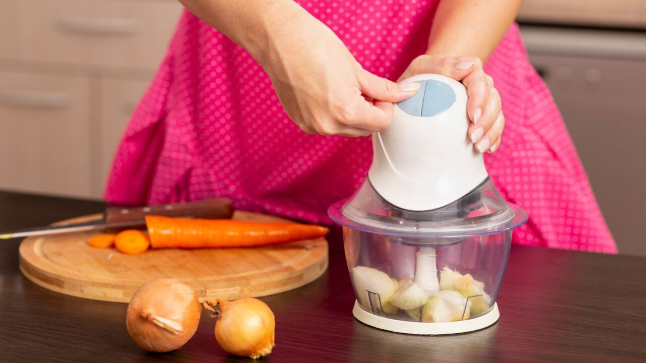 A woman chopping vegetables in a chopper placed on a wooden table.