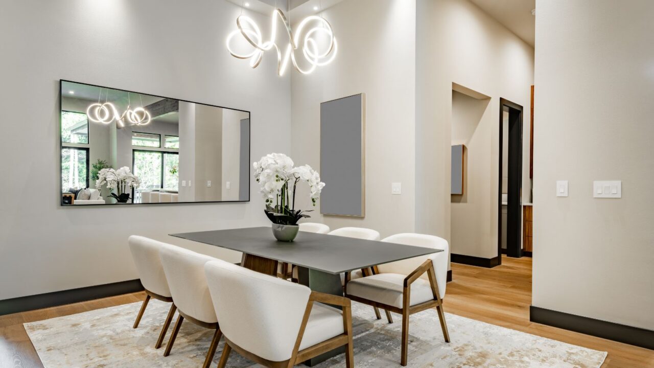 An elegant dining room interior with a dining table, chairs, flower vase, rug, a large mirror, and a chandelier hanging from the ceiling.