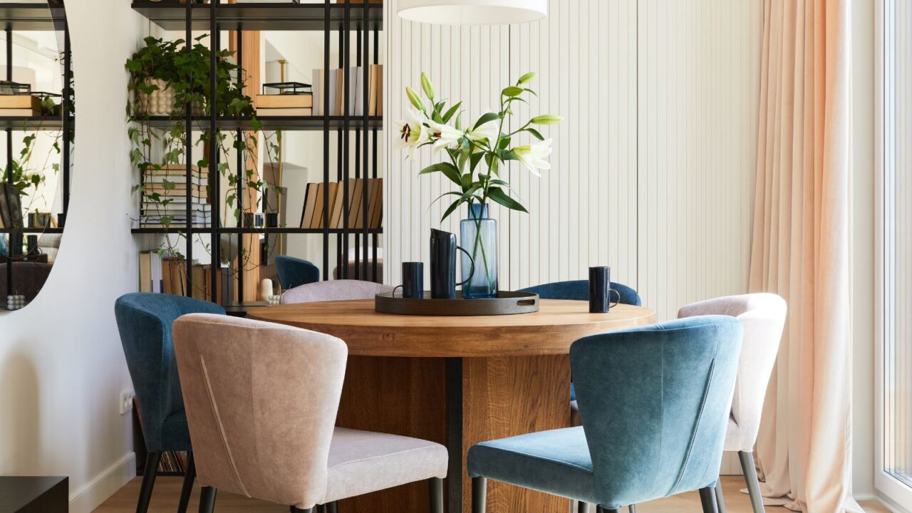 A stylish dining room interior with a wooden table, velvet chairs, bookshelf, mirror, and curtains.