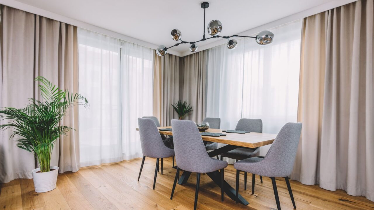 A stylish dining room interior with a wooden table, chairs, plant, curtains, and chandelier hanging from the ceiling.