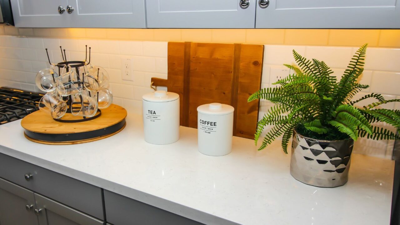 A kitchen counter with coffee and tea jars, lazy susan with glass cups, and a boston fern plant.