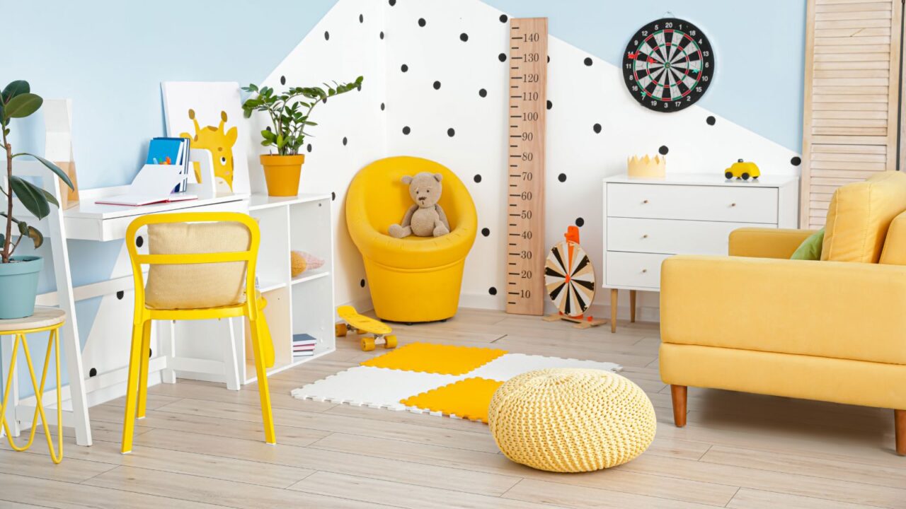 A vibrant playroom with mustard yellow accents including a cozy armchair, pouf, and chair, a rug, and a white desk with storage.