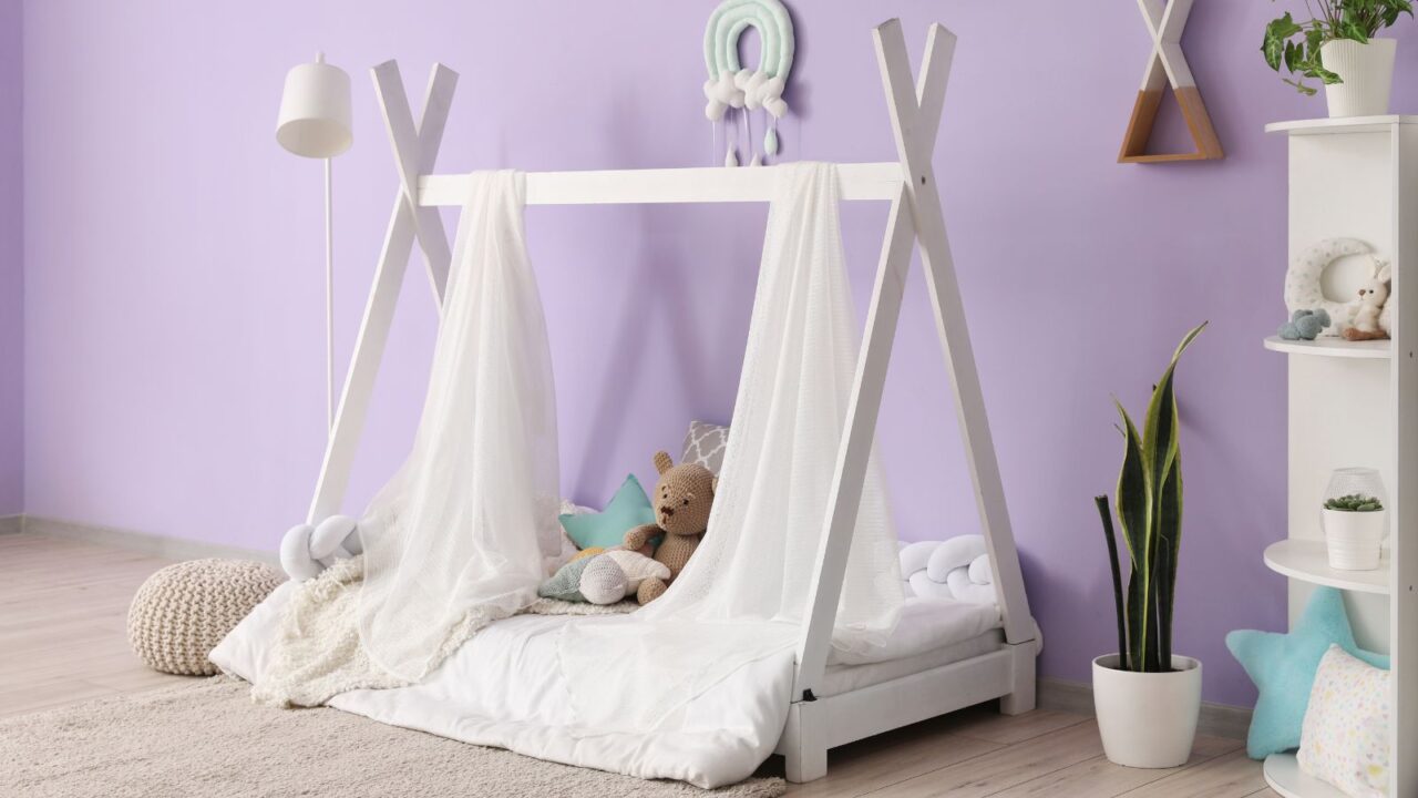 A lavender color playroom with a white bed, stuff toys, plant, and a white shelf with toys.