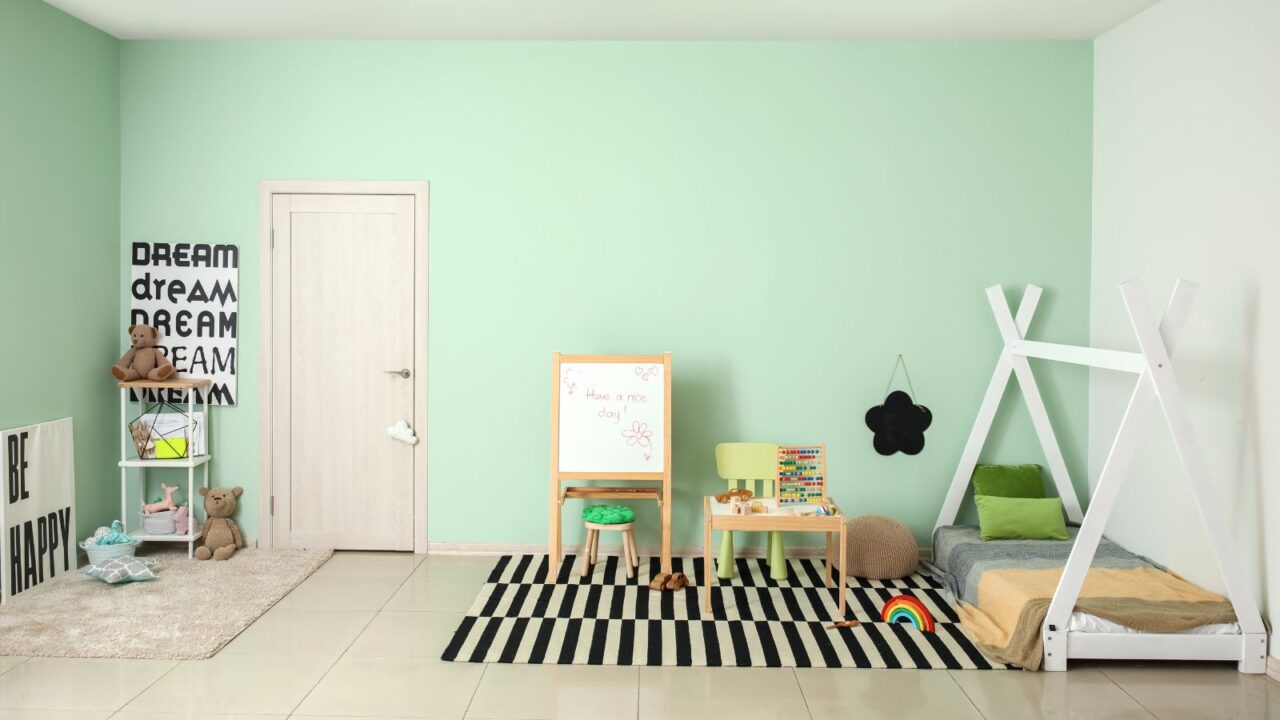 A stylish playroom with mint green walls, small table with toys, chalkboard easel, striped rug, and cozy decor elements.