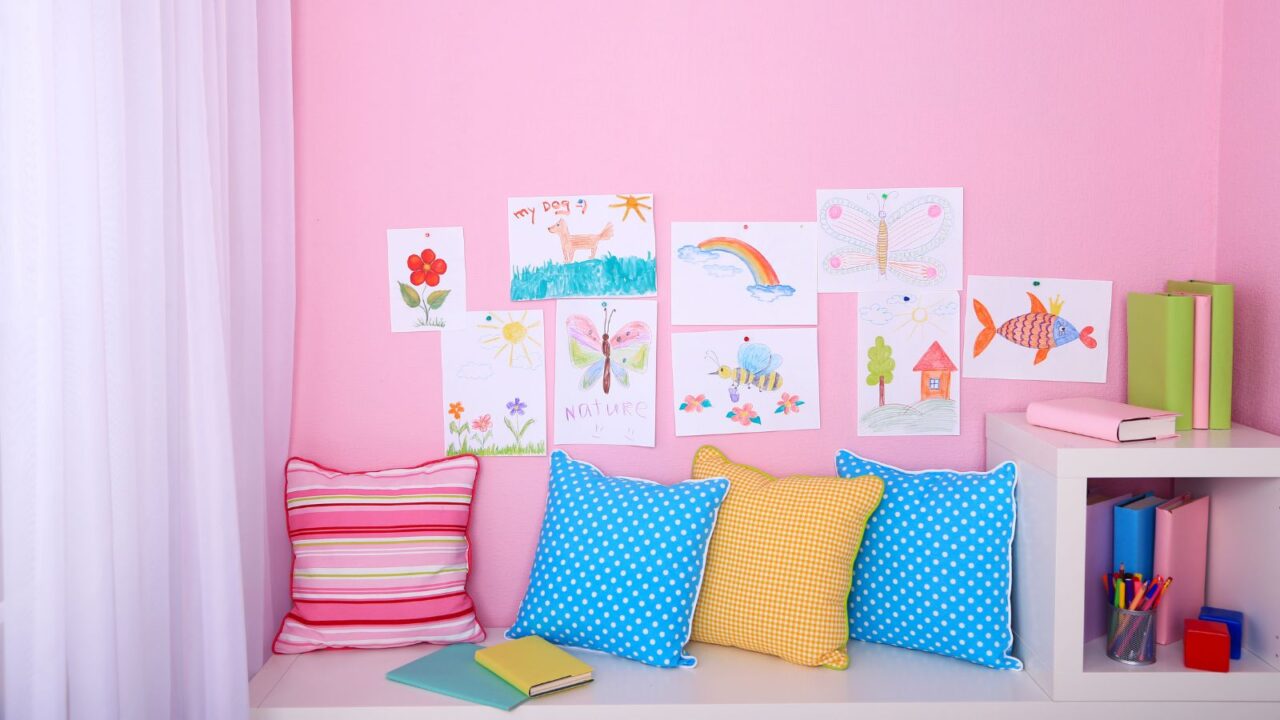 A playful room with a white bookshelf, colorful pillows, and drawings pasted on the pink wall.