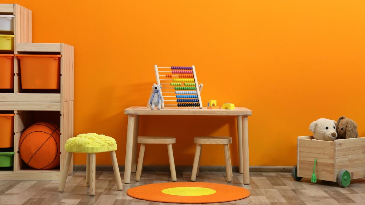 A playroom interior with a wooden table, chair, toy cart, stool, and a wooden shelf in front of a tangerine wall.