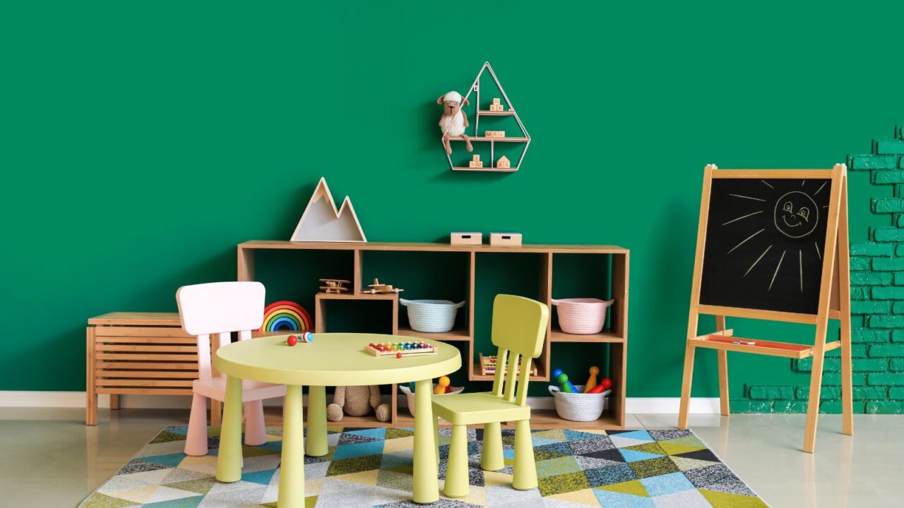 A playroom interior with a table, chair, drawing canvas, and a wooden shelf in front of an emerald green wall.