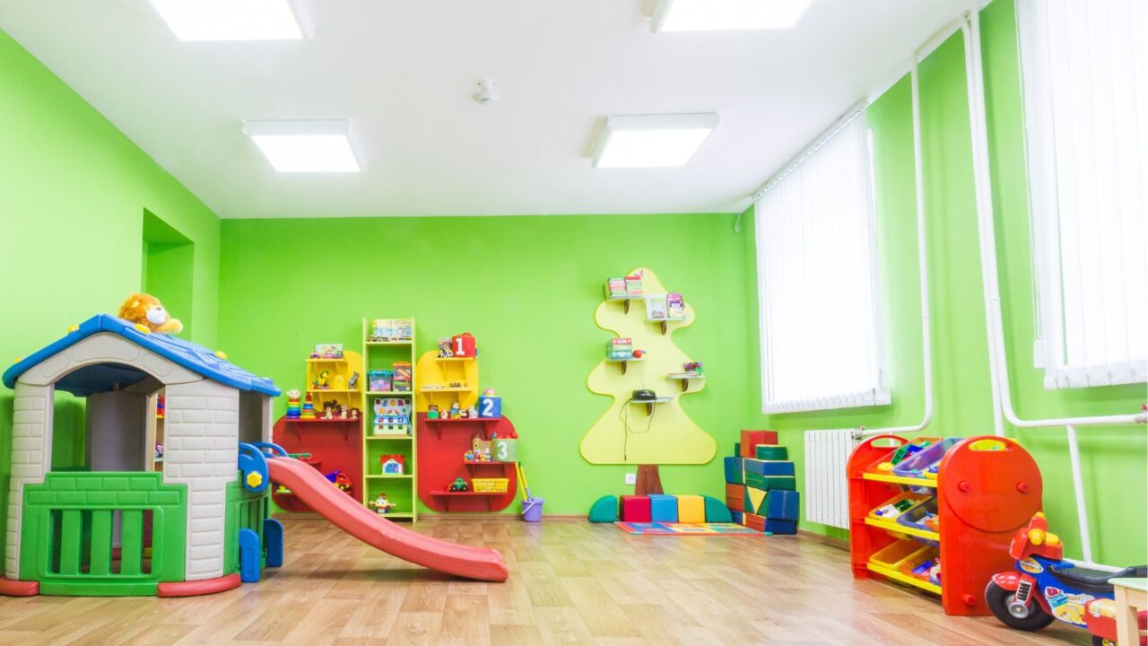 A children's playroom with bright green walls, colorful toys, a playhouse with a slide, and storage shelves.