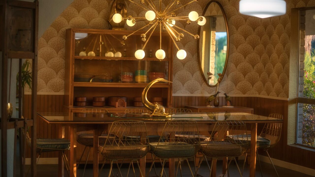 A dining room interior with metallic chairs, gold chandelier, glass table, shelf displaying dinnerware, and vintage-inspired decor accents.