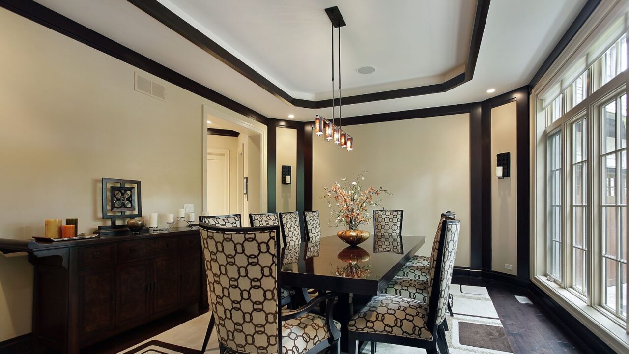 An elegant dining room interior with a recessed ceiling, dark wood furniture, patterned chairs, and pendant lighting.