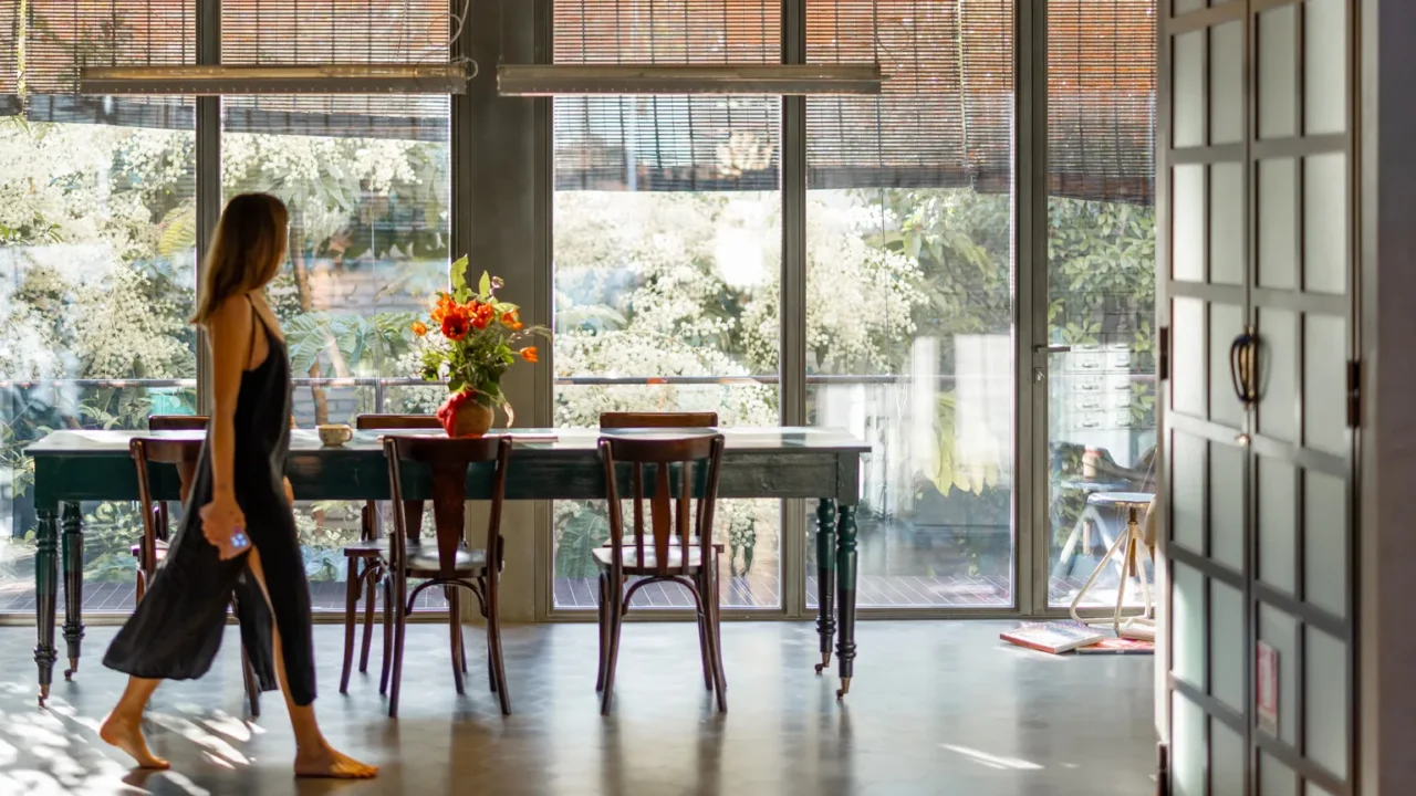 A woman in a black dress walks by a dining table with flowers and large windows.