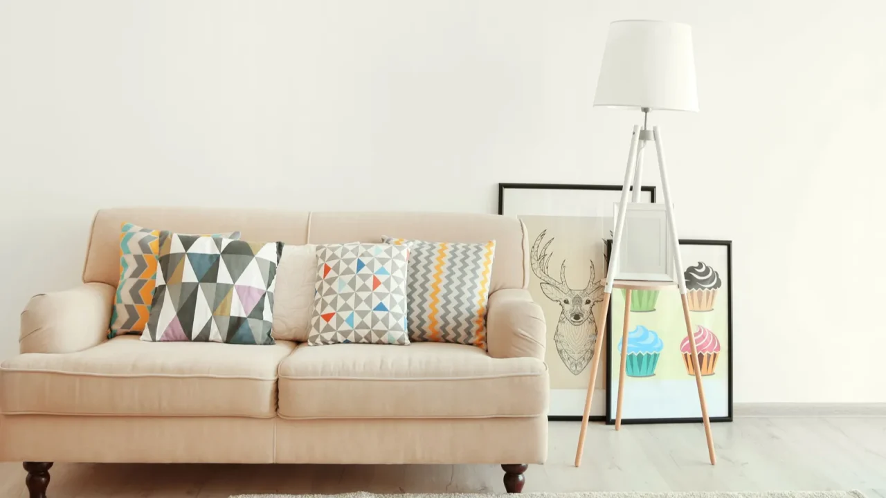 A stylish living room interior with a floor lamp, art frames, and a beige sofa with geometric patterned throw pillows on it.