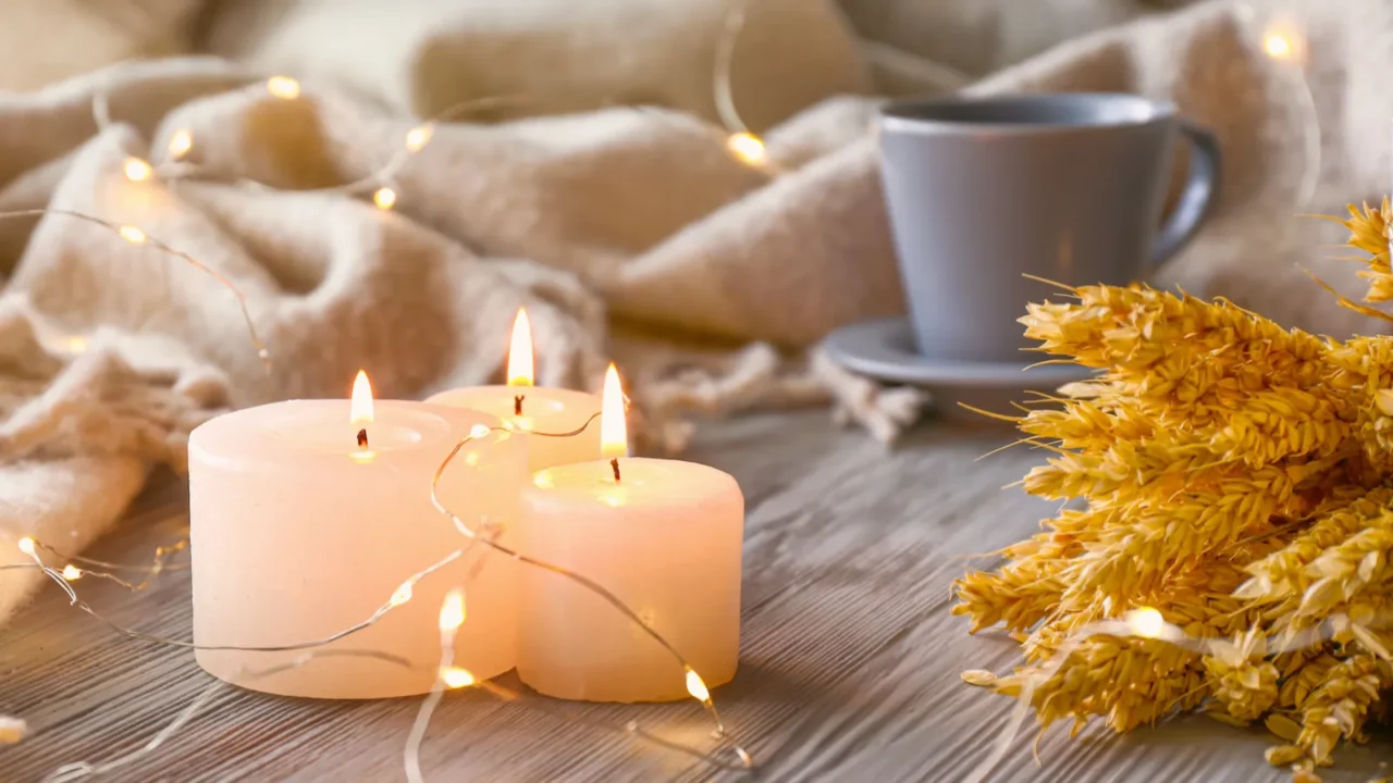 Candles and a coffee cup beside wheat decor and fairy lights on a soft blanket.