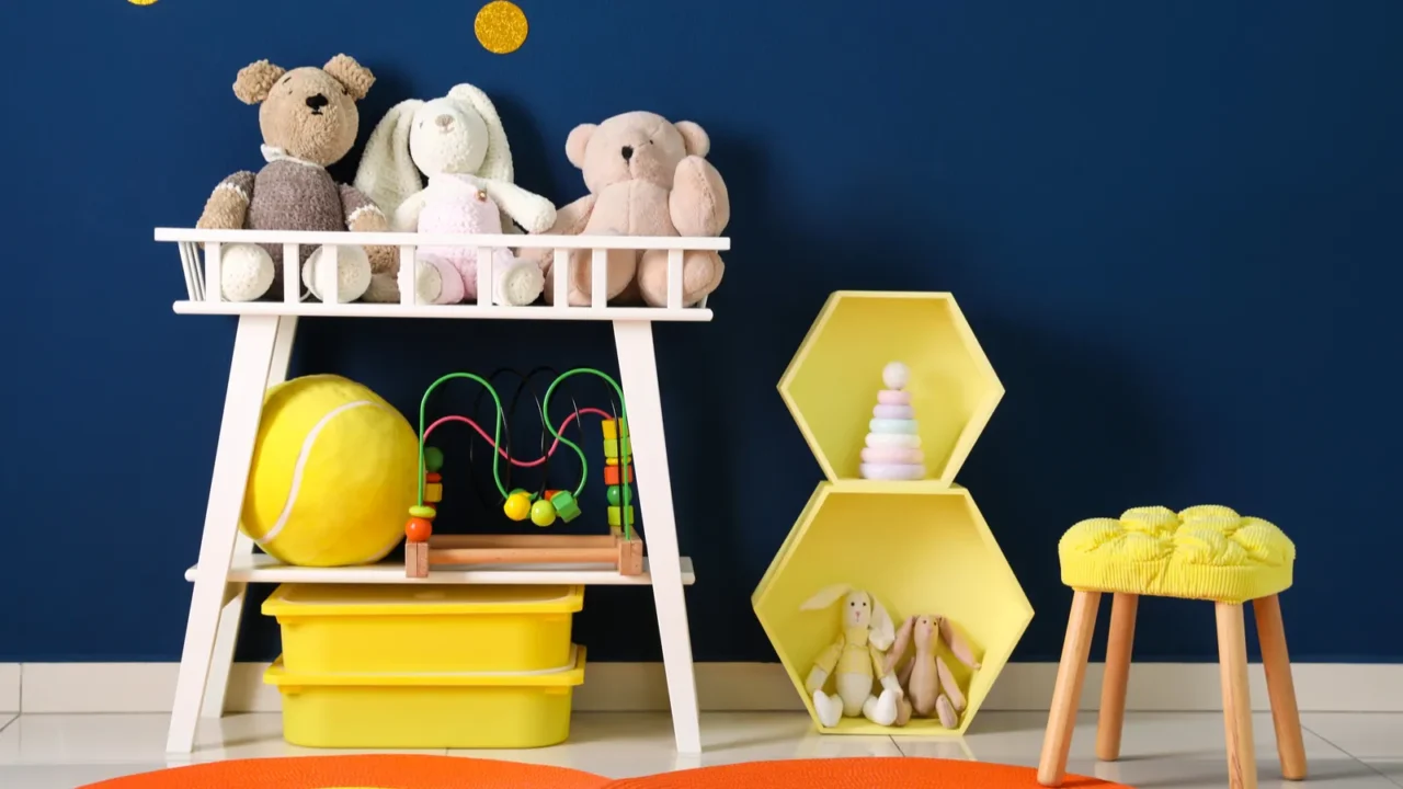 A playroom with a yellow stool, orange rug, yellow shelves with toys, and a white toy shelf in front of a navy blue wall.