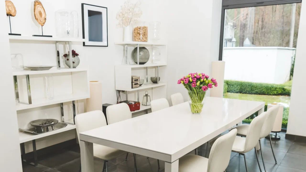 A dining room interior with a white dining table, chairs, flower vase, and a feature wall with open shelves with decor accents.