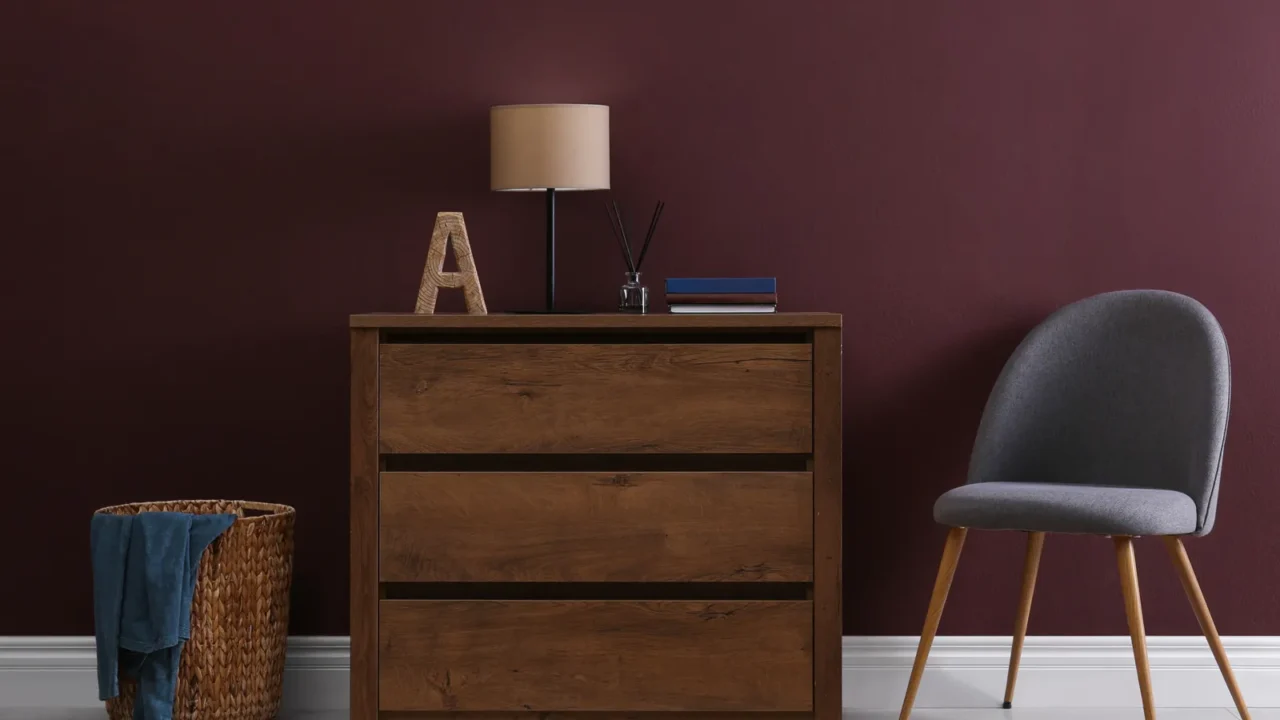 A wooden dresser with a lamp and an ornamental letter beside a gray chair against a rich burgundy wall.