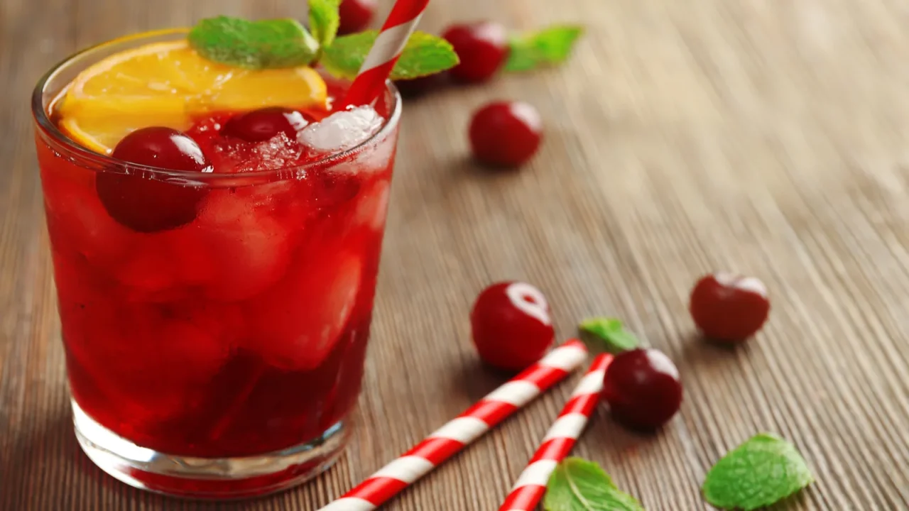 A glass of cherry drink garnished with lemon slices and mint leaves, surrounded by cherries.