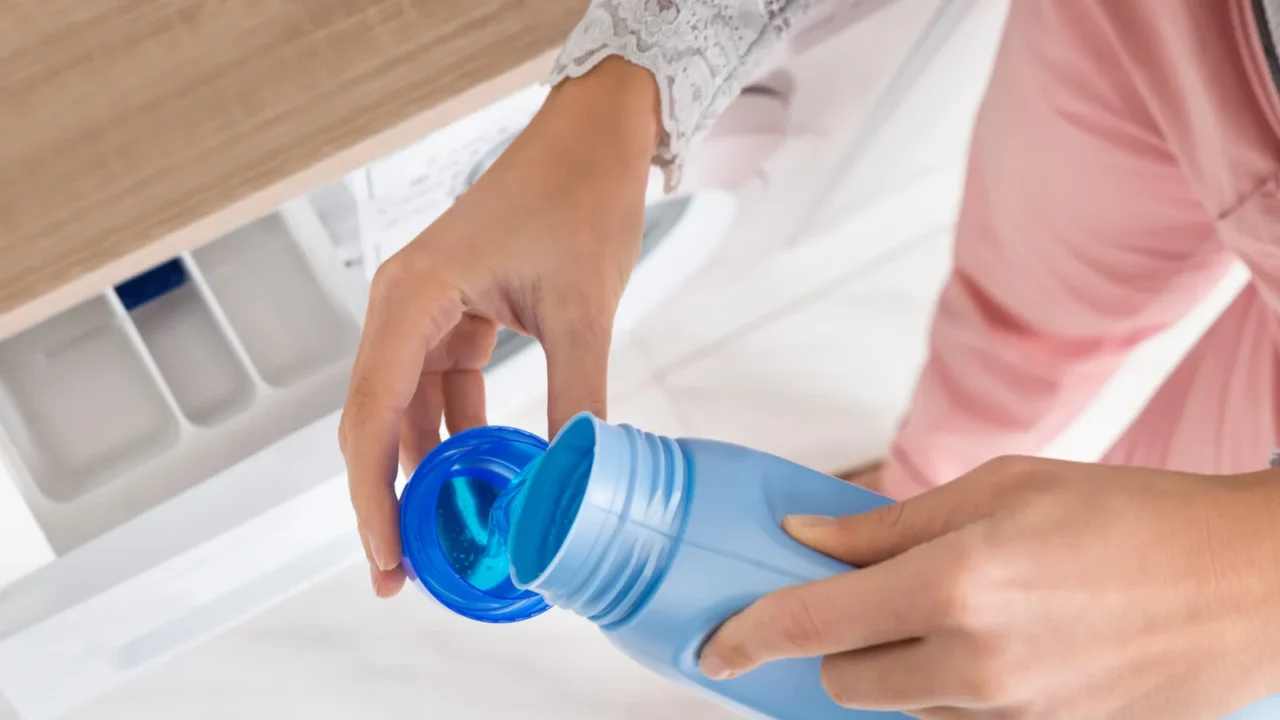 A hand pouring blue detergent into a washing machine compartment.