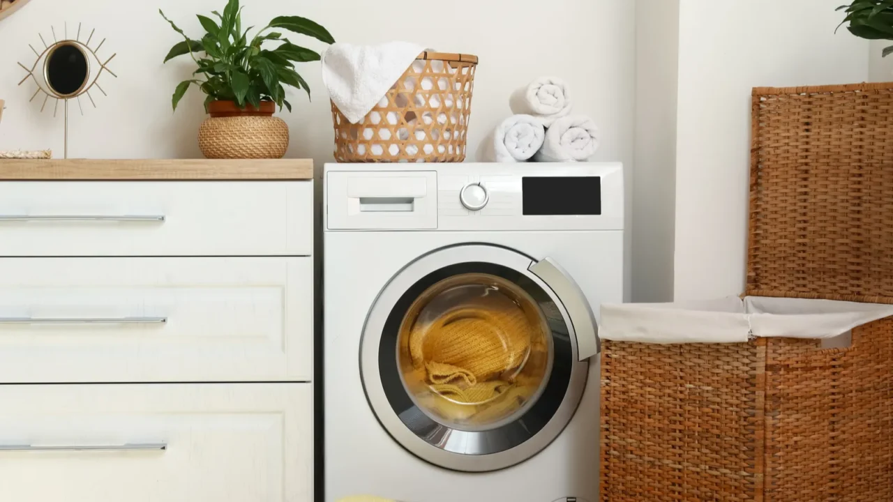 A modern laundry room featuring a washing machine, wicker basket, folded towels, and a potted plant.