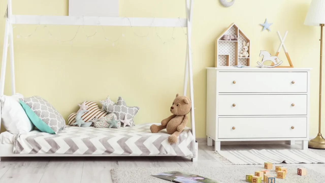 A playroom interior with with soft buttercream yellow walls, a low white canopy bed with throw pillows and stuff toy, a rug, a white dresser with decorative accents, wood toys, and a floor lamp.