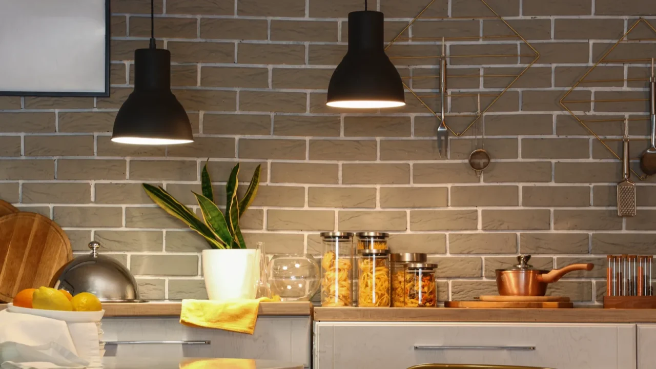 A modern kitchen scene featuring pendant lights, potted plants, and various utensils against a brick wall.