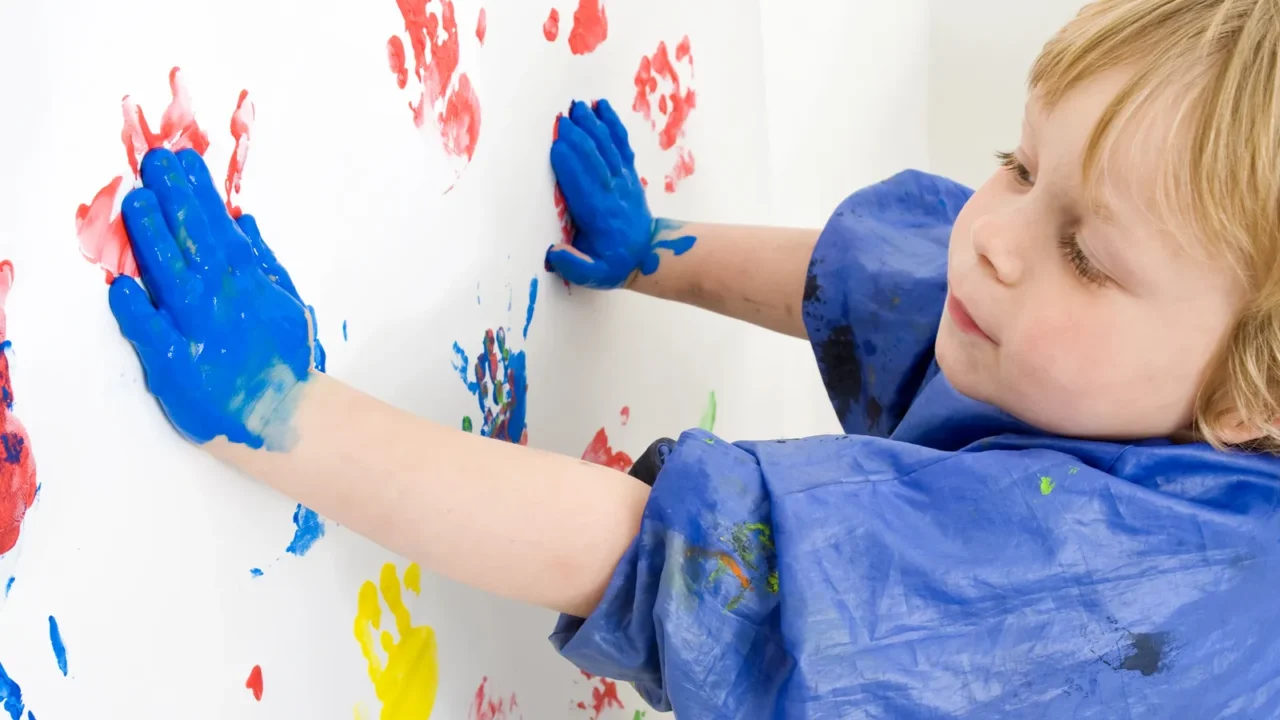 A little boy doing hand painting on a white wall.